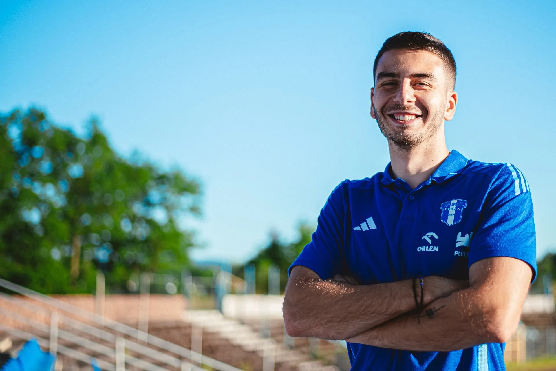Na zdjęciu widać uśmiechniętego mężczyznę w niebieskiej koszulce sportowej, stojącego na tle jasnego nieba i zieleni.