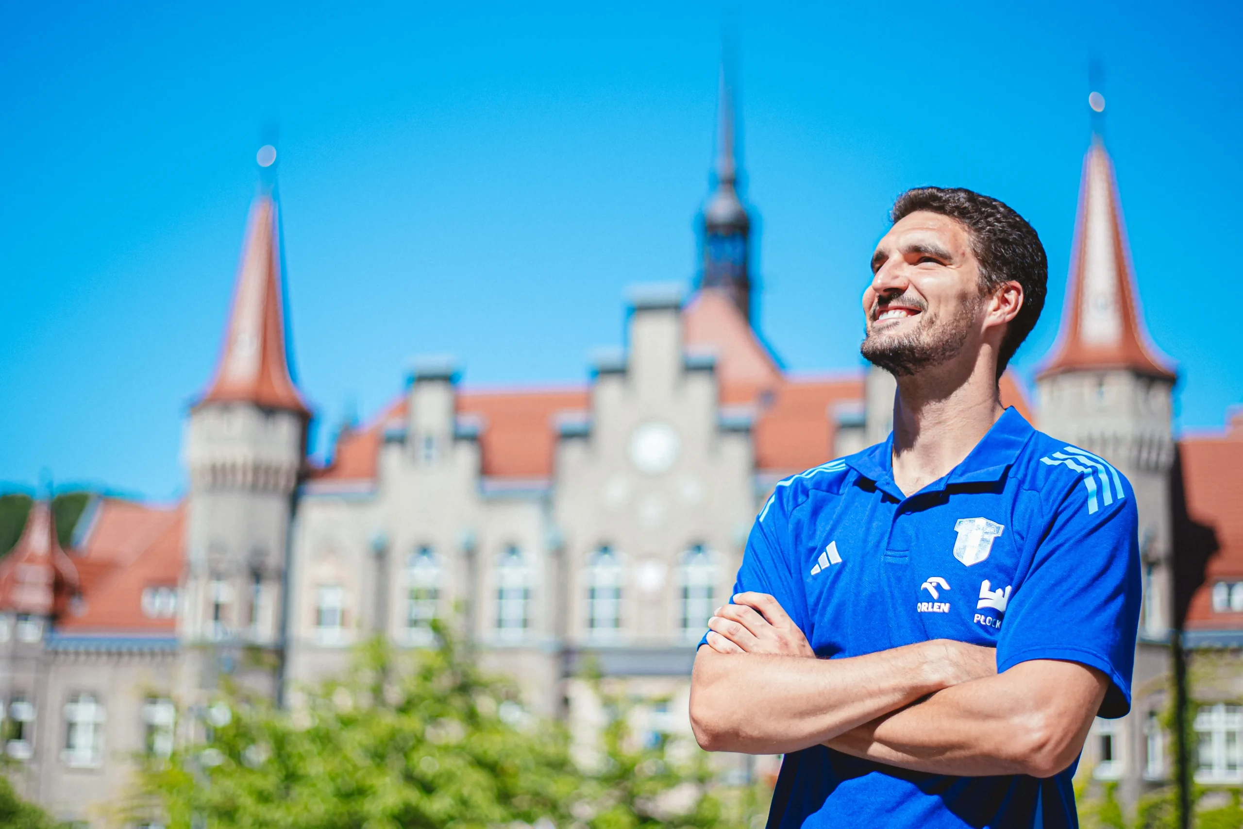 Mężczyzna w niebieskiej koszulce sportowej z uśmiechem stoi na tle zabytkowego budynku z wieżyczkami na jasnym niebie.