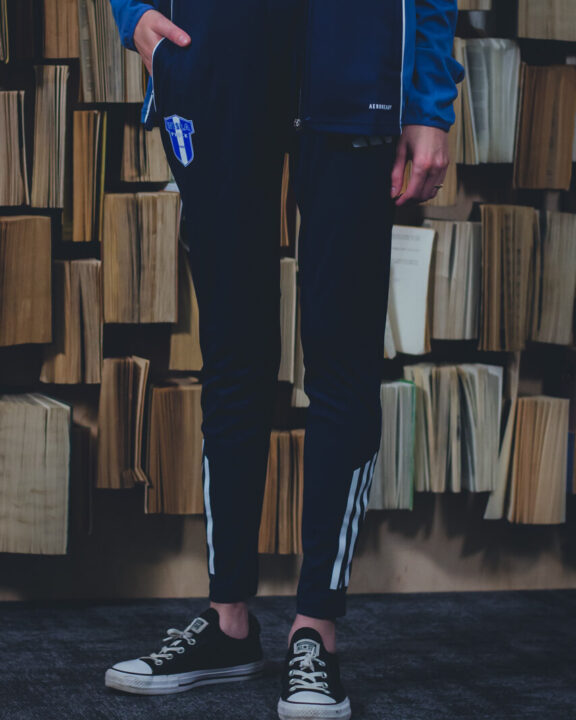 Osoba stojąca w granatowych spodniach Adidas 25/26 XXL z białymi paskami po bokach, na czarnych sneakersach, na tle regału z książkami.