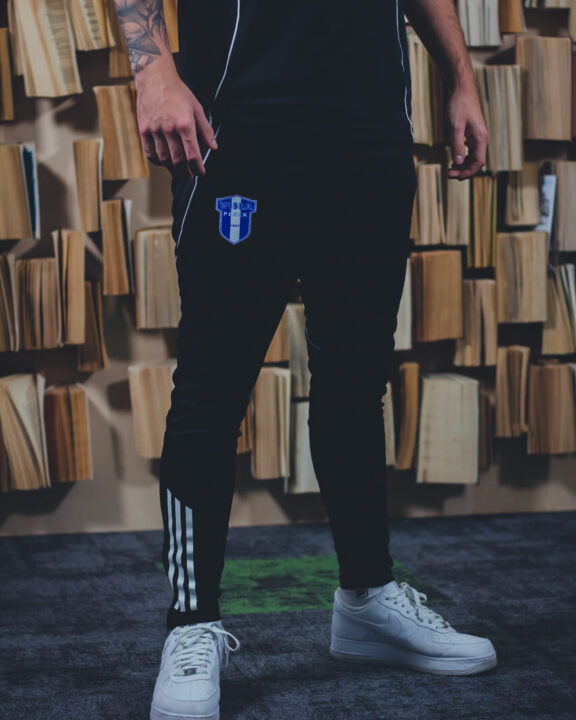 Osoba stojąca w czarnych spodniach Adidas 25/26 XXL z białymi paskami i naszywką Wisła Płock na udzie, w białych sneakersach, na tle regałów z książkami.