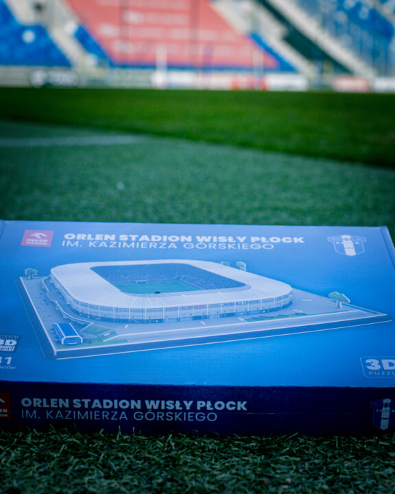 Niebieskie pudełko puzzli 3D z wizerunkiem stadionu Orlen Stadion Wisły Płock im. Kazimierza Górskiego leżące na trawie boiska.