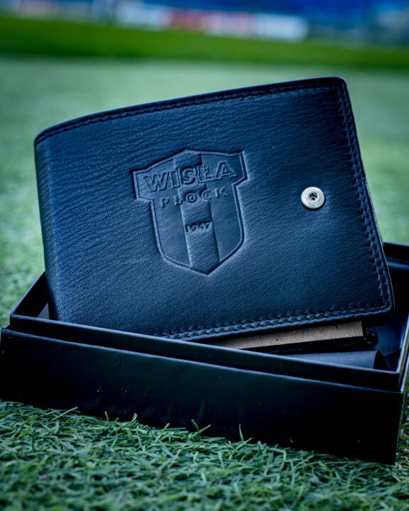 Czarny skórzany portfel męski z wytłaczanym godłem Wisły Płock, leżący na czarnym pudełku na zielonej trawie.