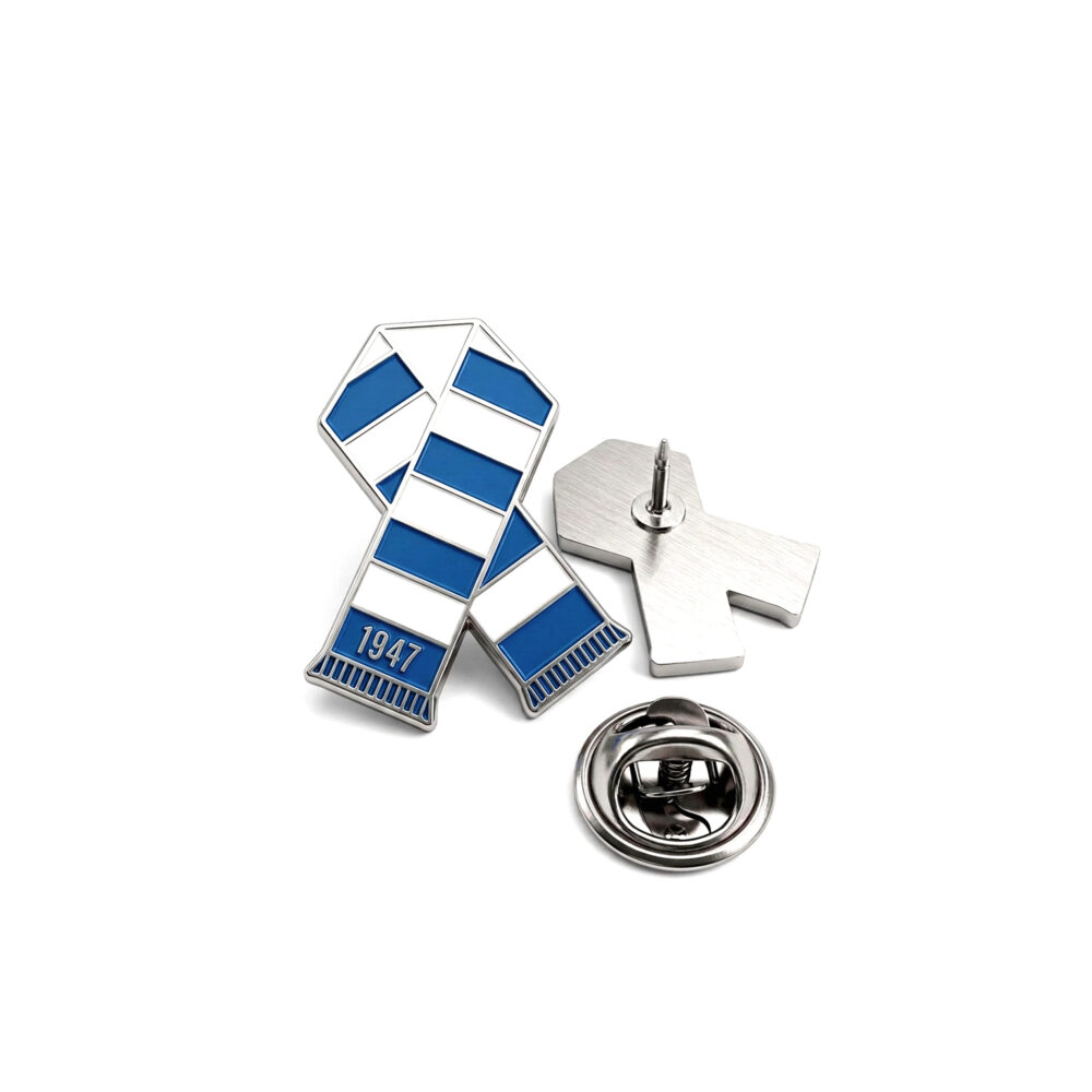 Srebrny pin w kształcie stylizowanego szalika z niebiesko-białymi paskami i napisem 1947, leżący obok metalowego zapięcia.