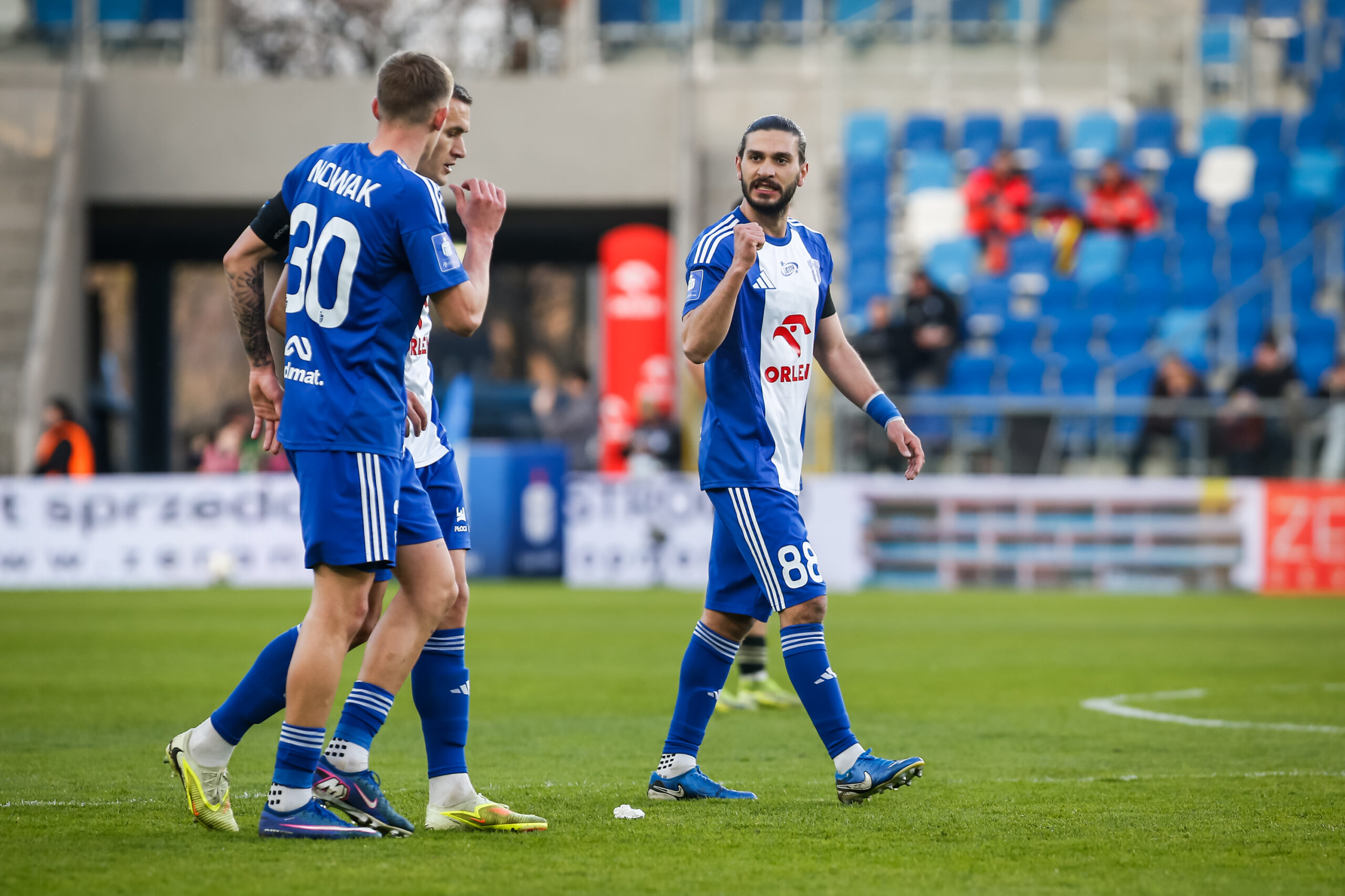 Trzech piłkarzy w niebiesko-białych strojach na boisku piłkarskim, jeden z nich unosi pięść w geście triumfu, a w tle widać trybuny.