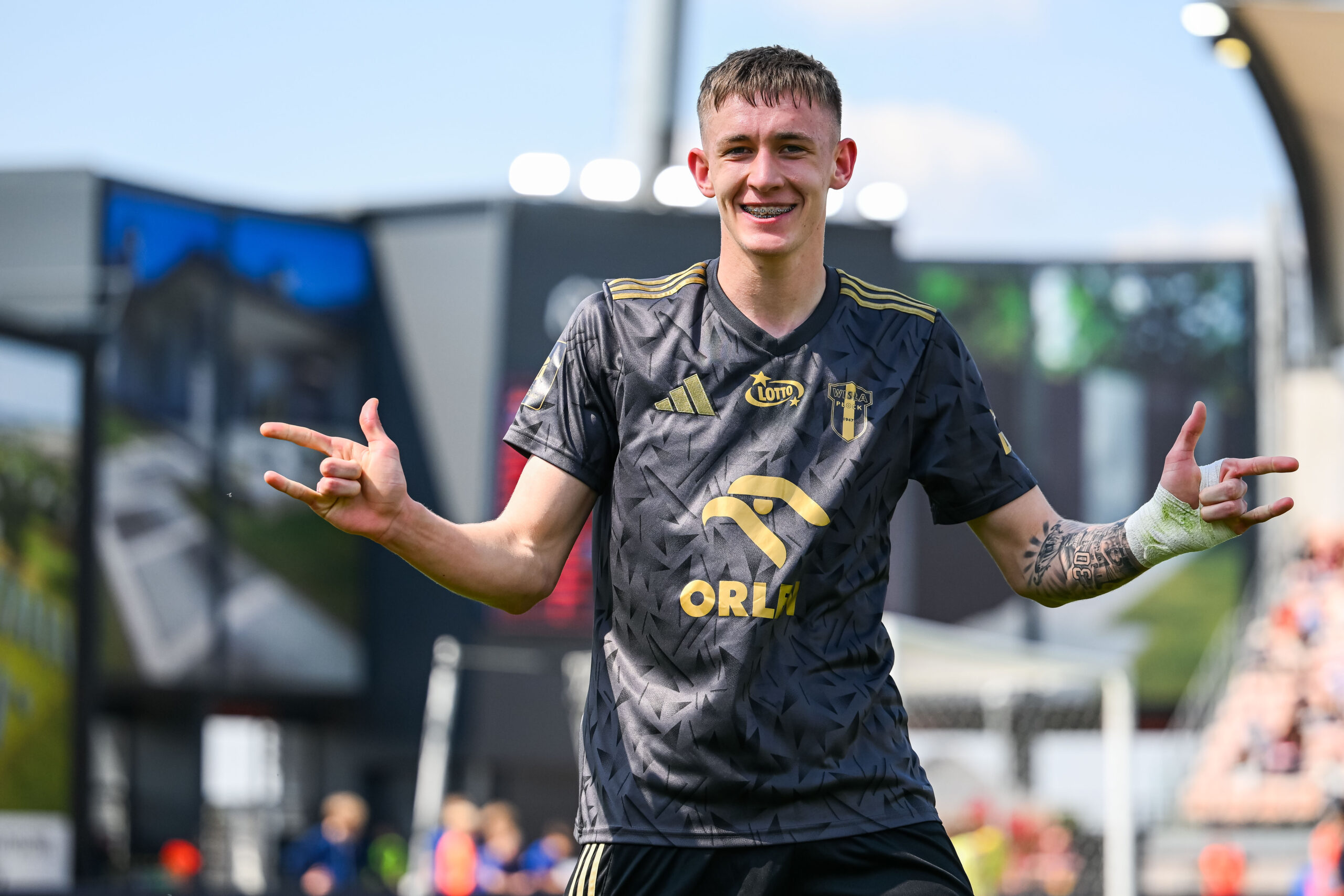 Piłkarz Wisły Płock w ciemnym stroju meczowym z żółtymi akcentami, uśmiechnięty, wykonuje gesty rękami podczas meczu.