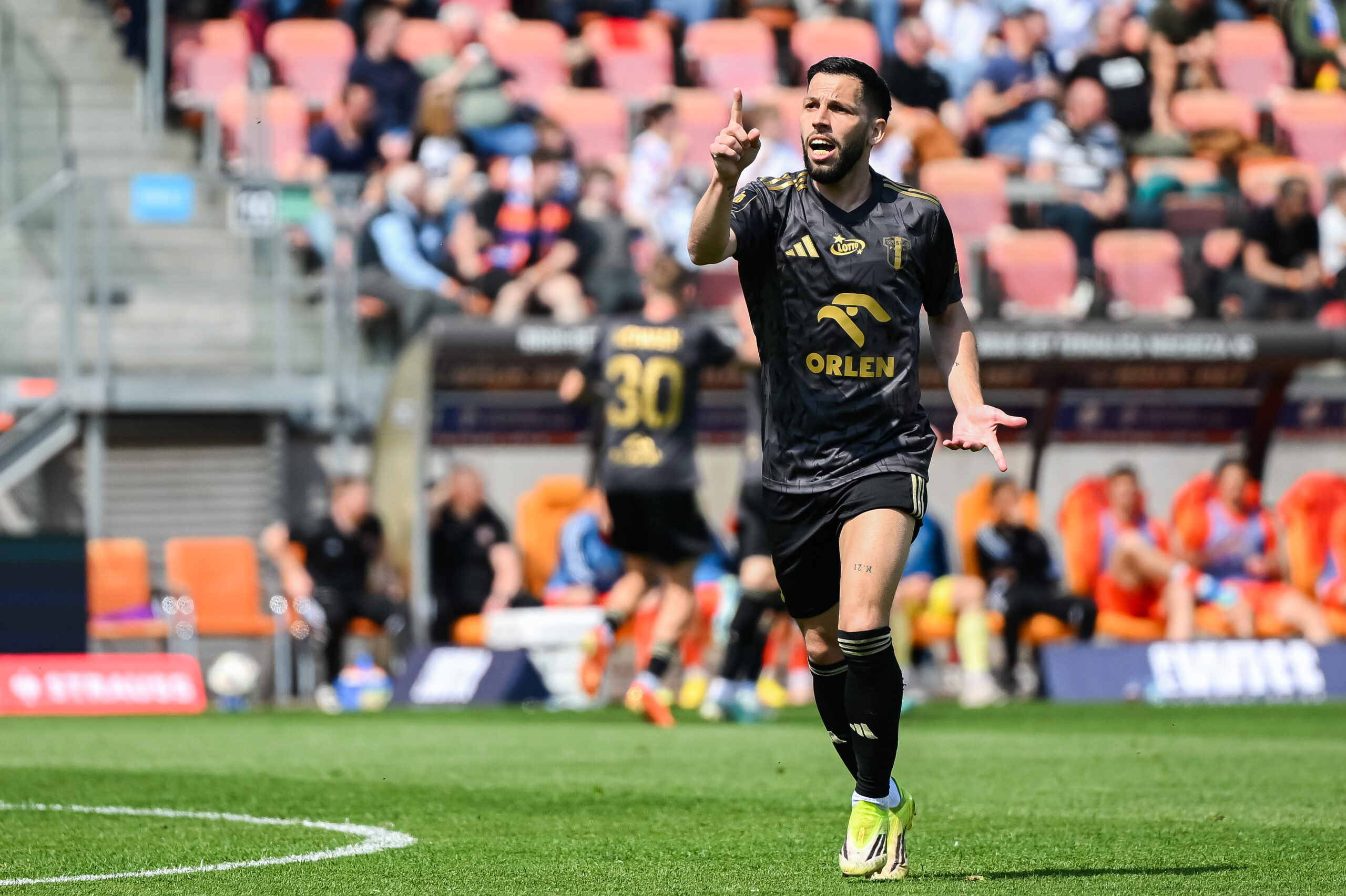 Piłkarz w czarnym stroju z żółym logo Orlen celebruje na boisku, wskazując palcem ku górze, w tle kibice na trybunach.