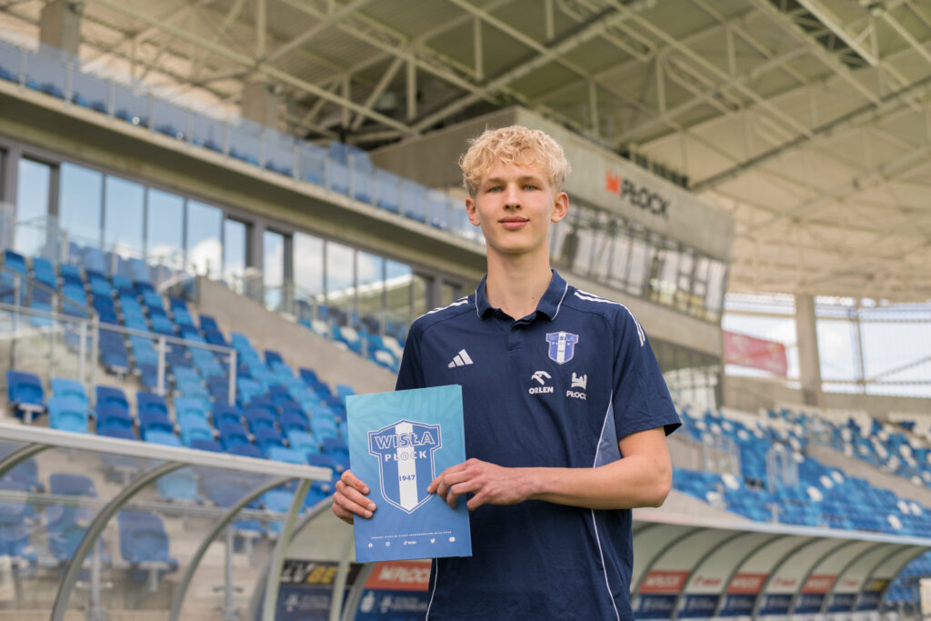 Młody mężczyzna w granatowej koszulce z herbem Wisły Płock stoi na stadionie i trzyma folder z logiem klubu, sugerujący podpisanie profesjonalnego kontraktu.
