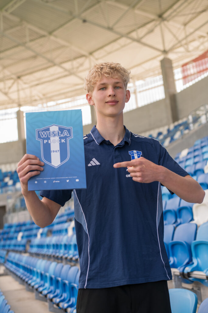 Młody piłkarz w klubowej koszulce Wisły Płock prezentuje podpisany kontrakt, trzymając niebieski folder z logo klubu na tle stadionowych trybun.