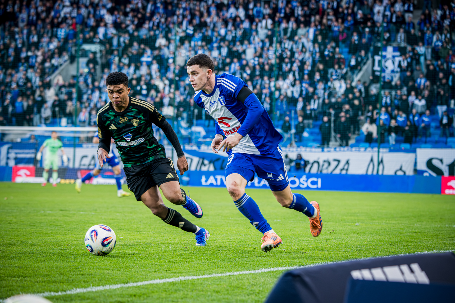 Dwóch piłkarzy rywalizuje o piłkę na boisku podczas meczu, przy tłumnie zgromadzonych kibicach na trybunach.