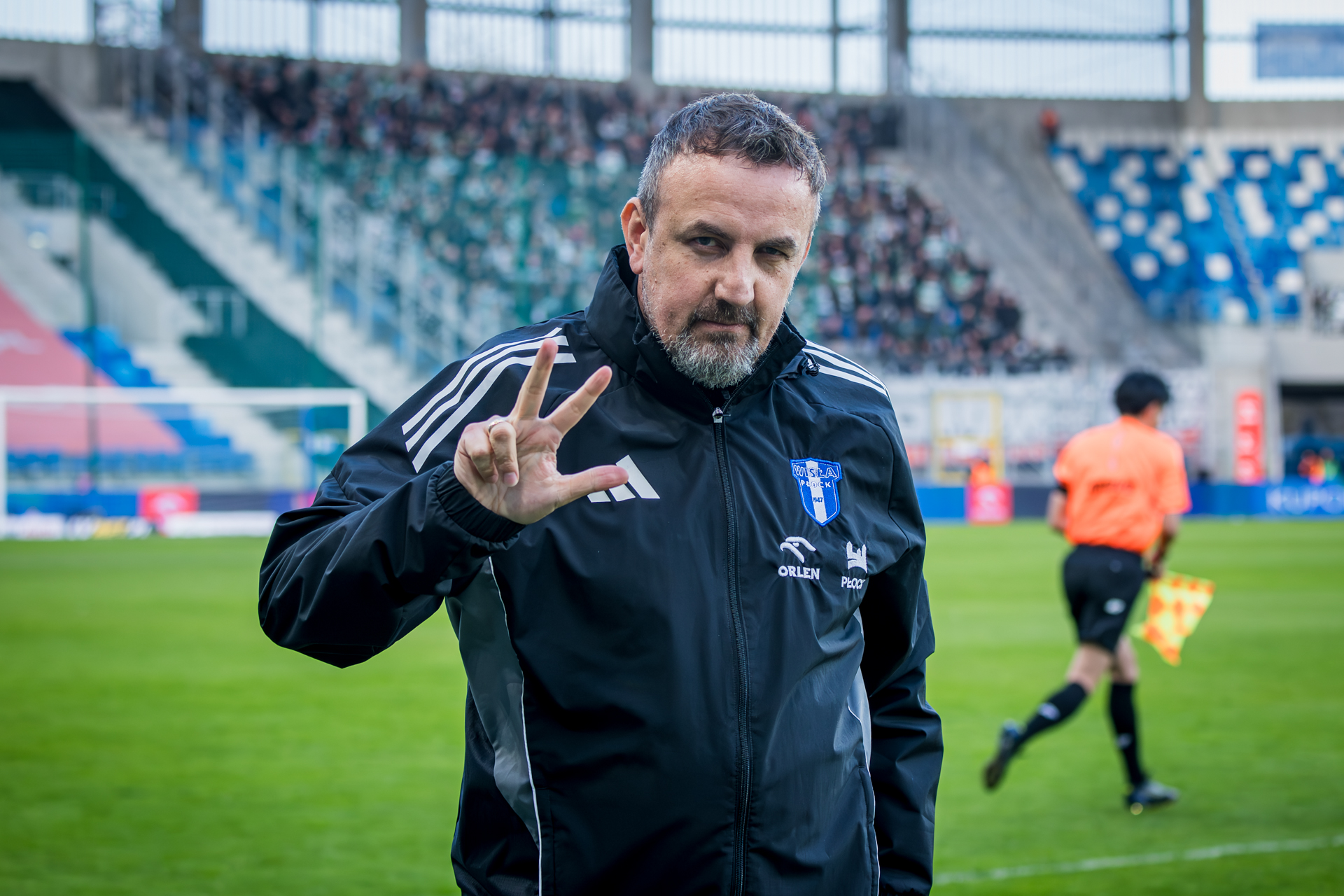Trener piłkarski w czarnej kurtce z logo klubu stoi na boisku przed trybunami i pokazuje gest dłoni w stronę kamery.