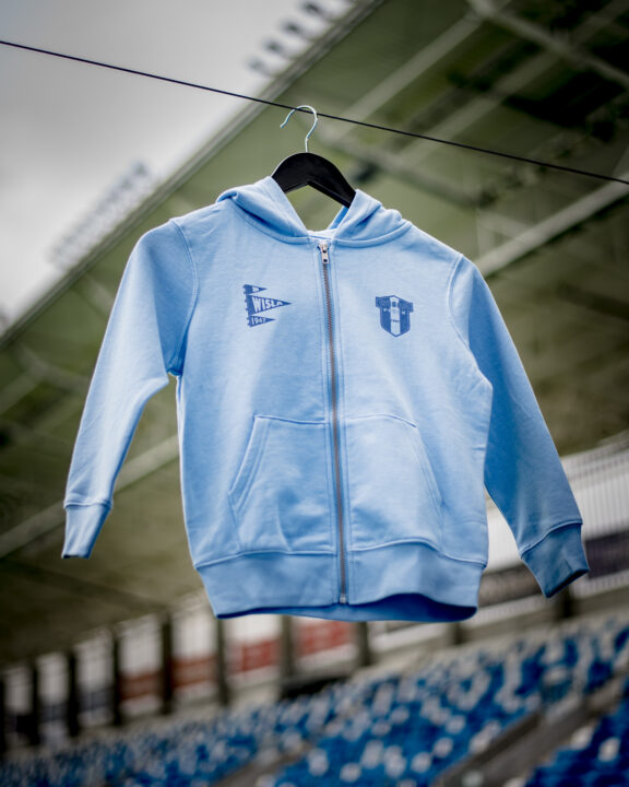 Niebieska dziecięca bluza z kapturem i zapięciem na suwak, zawieszona na czarnym wieszaku na tle stadionowych trybun z niebieskimi siedzeniami, z logo Wisła Płock na piersi.
