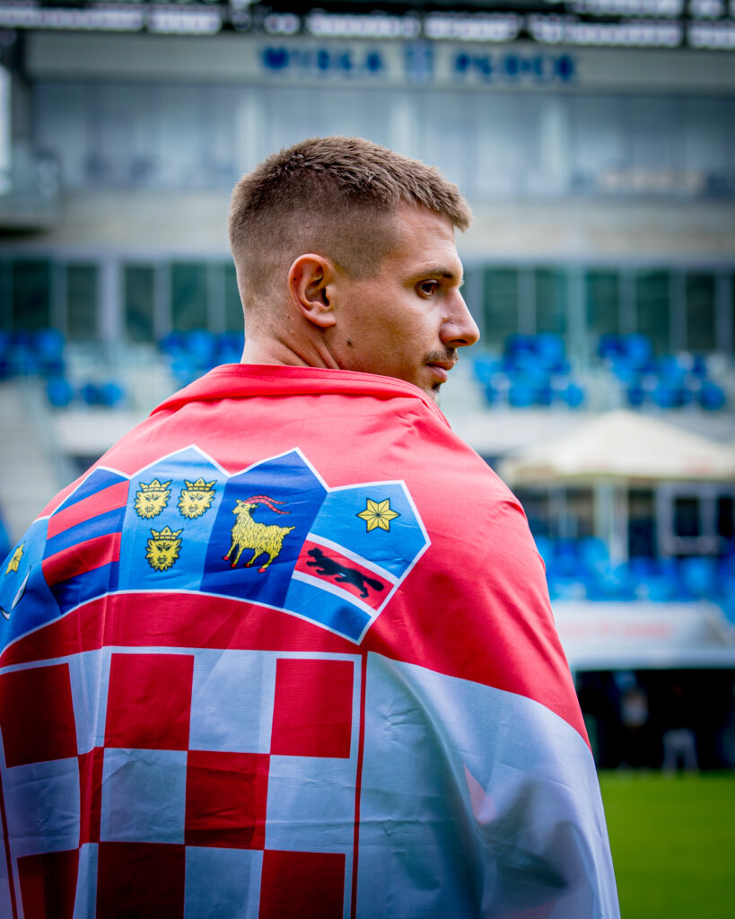 Mężczyzna widziany z profilu, ubrany w czerwoną koszulkę z kolorowym herbem na plecach, stoi na tle stadionu z niebieskimi krzesełkami.