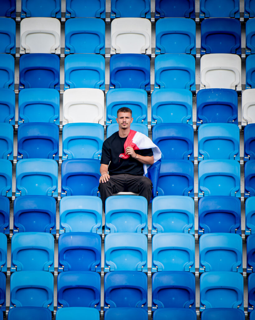 Mężczyzna siedzi na tle rzędów niebieskich i białych krzeseł stadionowych, trzymając polską flagę.