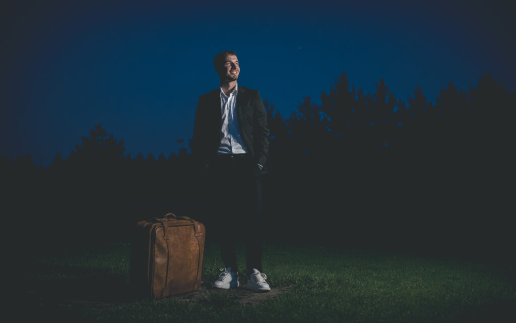 Mężczyzna w garniturze stoi na trawie wieczorem z dużą brązową walizką, uśmiechnięty, wraca do domu na tle nocnego nieba i drzew.
