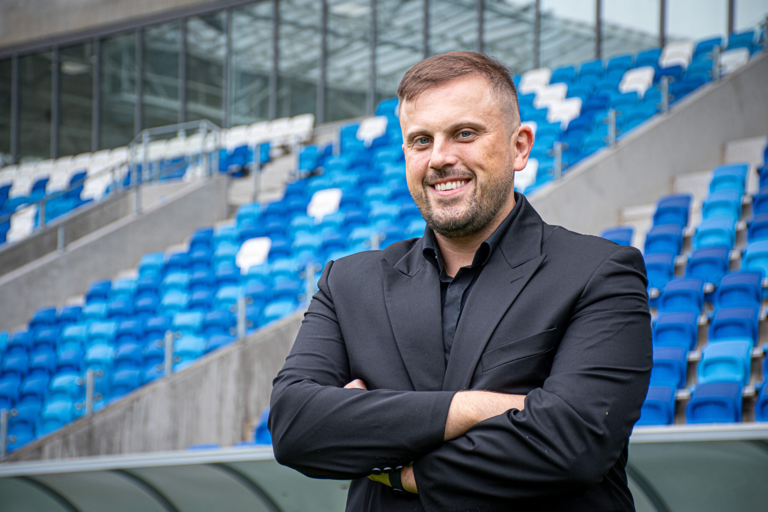 Mężczyzna w czarnym garniturze na tle stadionowych trybun, będący dyrektorem sportowym Wisły Płock S.A.