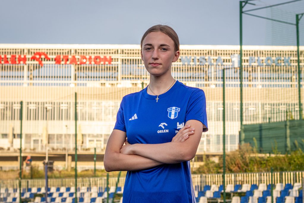 Młoda zawodniczka w niebieskiej koszulce sportowej stoi na boisku z założonymi rękoma, na tle ogrodzenia i trybun.