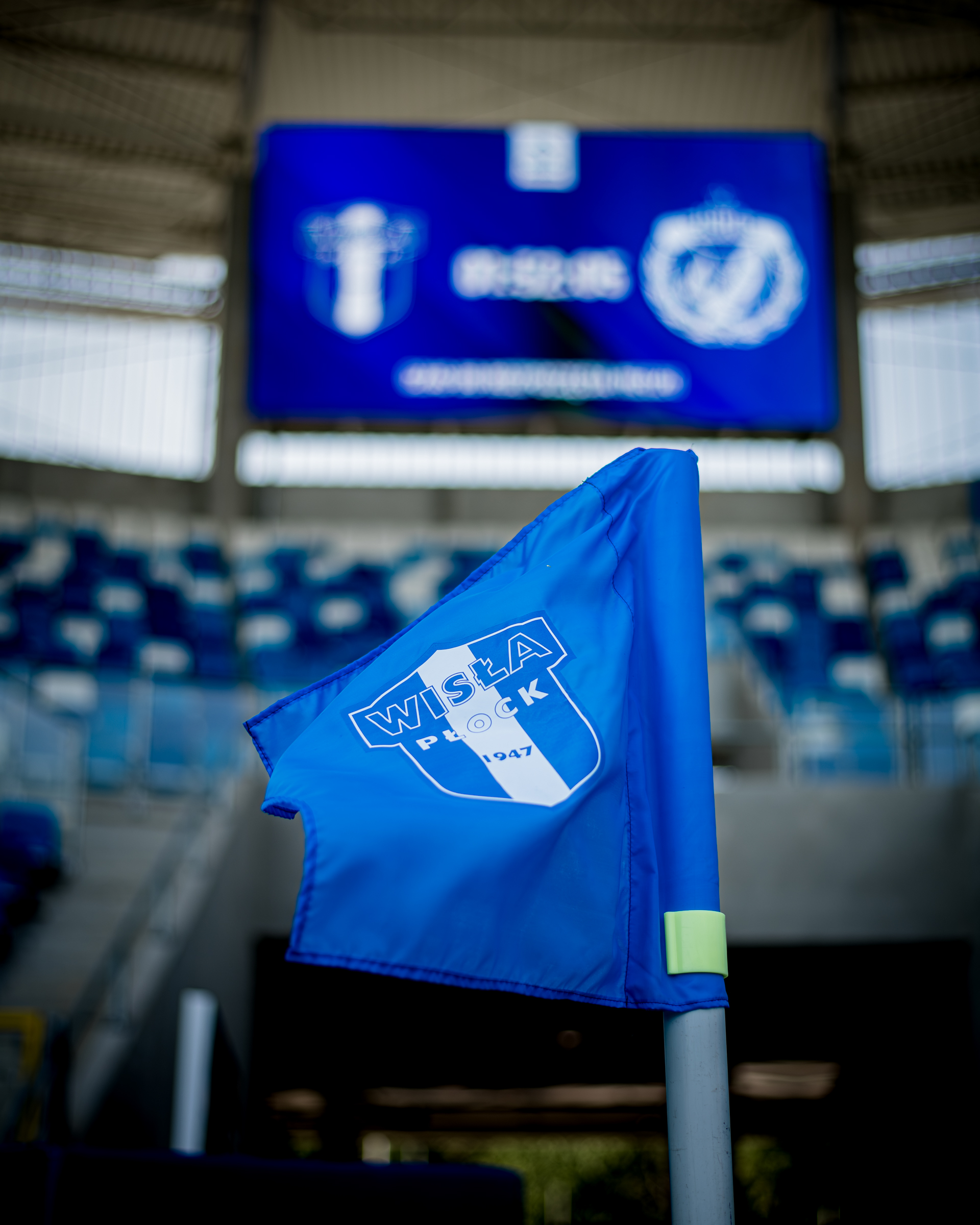 Flaga Wisły Płock z herbem na niebieskim tle, ustawiona przed trybunami stadionu.