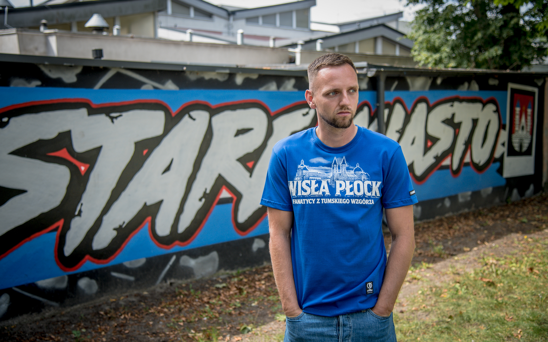Mężczyzna w niebieskiej koszulce z napisem Wisła Płock stoi na tle kolorowego graffiti na murze.