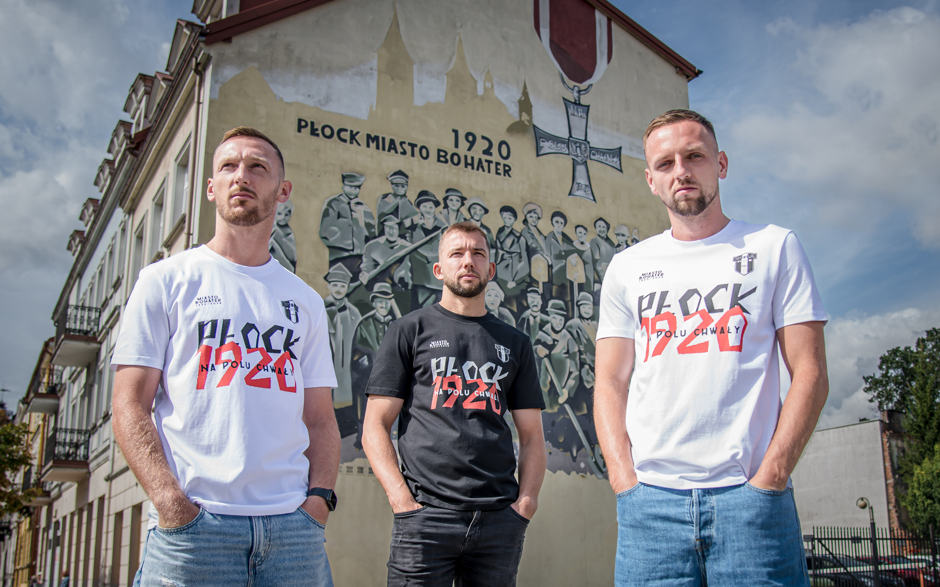 Trzech mężczyzn stoi przed muralem upamiętniającym bohaterów Płocka z napisem 1920 – Płock Miasto Bohater.
