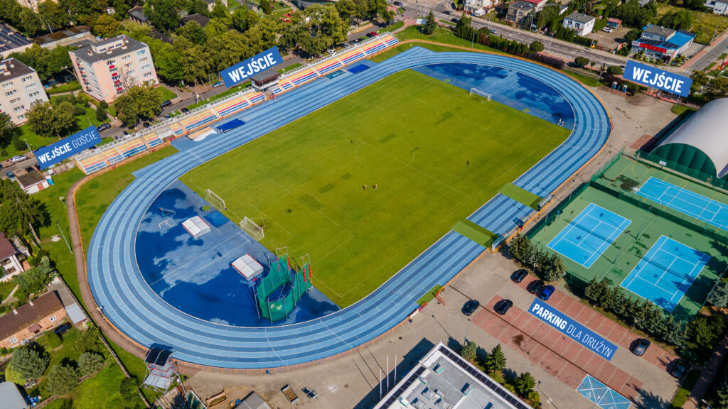 Widok z lotu ptaka na stadion lekkoatletyczny z niebieską bieżnią wokół zielonego boiska i oznaczeniami WEJŚCIE wskazującymi wejścia na mecz Wisła II Płock – Lechia Tomaszów Mazowiecki.
