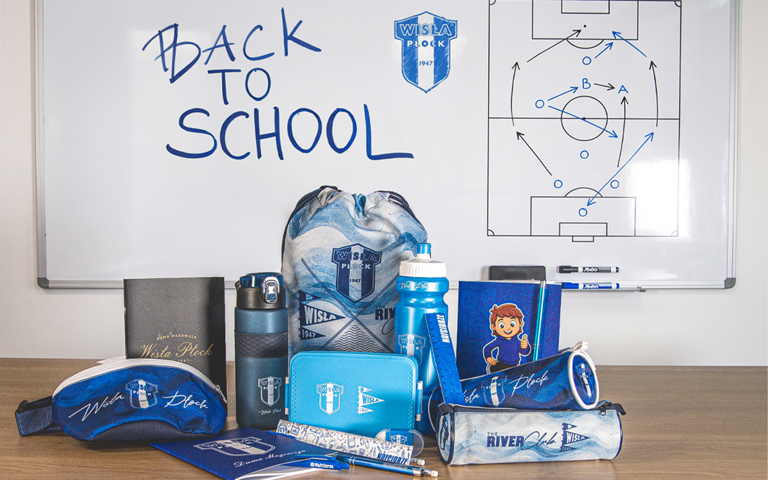 Zestaw szkolnych przyborów w niebieskich odcieniach (plecaki, piórniki, bidony) ustawiony na biurku przed tablicą z napisem „Back to School” i logo Wisły Płock.